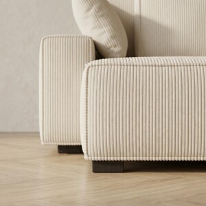 Close-up detail of a cream corduroy modular sofa showing the thick square armrest, wide chaise edge, and vertical ribbed upholstery texture. The image focuses on the tailored piping, soft fabric finish, and dark wooden block feet resting on pale herringbone flooring. A matching throw pillow and back cushion appear softly in the background, highlighting the sofa’s premium construction, clean seam work, and modern minimalist design for stylish living room interiors.