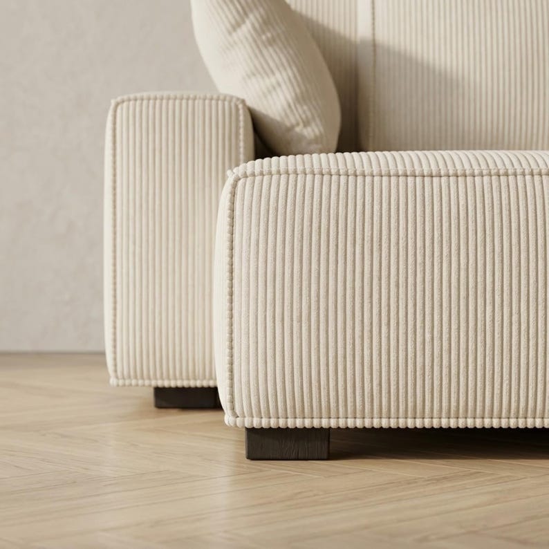 Close-up detail of a cream corduroy modular sofa showing the thick square armrest, wide chaise edge, and vertical ribbed upholstery texture. The image focuses on the tailored piping, soft fabric finish, and dark wooden block feet resting on pale herringbone flooring. A matching throw pillow and back cushion appear softly in the background, highlighting the sofa’s premium construction, clean seam work, and modern minimalist design for stylish living room interiors.