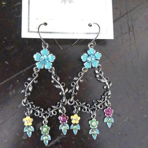 May include: A pair of dangle earrings featuring a dark metal vine-like frame with a turquoise flower at the top. The bottom of each earring is adorned with small yellow, pink, and green enameled flower charms. The earrings are attached to a CSD Design card.