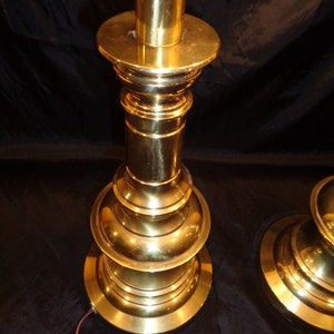 May include: A pair of vintage brass table lamps. Each lamp features a tiered base and a cylindrical column, all in a polished gold finish. The lamps have a classic, elegant design, with a power cord extending from the base.