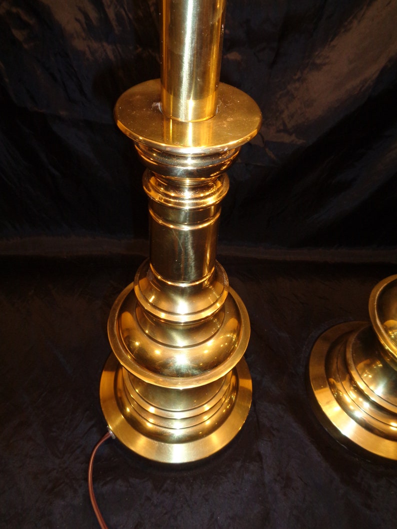 May include: A pair of vintage brass table lamps. Each lamp features a tiered base and a cylindrical column, all in a polished gold finish. The lamps have a classic, elegant design, with a power cord extending from the base.