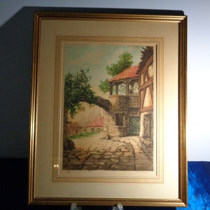 May include: Framed print of a European village scene. The artwork depicts a cobblestone street, stone buildings, and a bridge. The print is in a gold frame with a cream-colored mat.