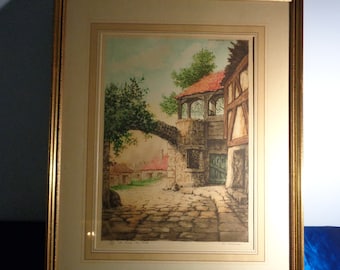 Pode incluir: Impressão emoldurada de uma cena de vila europeia. A obra de arte retrata uma rua de paralelepípedos, edifícios de pedra e uma ponte. A impressão está em uma moldura dourada com um passe-partout creme.