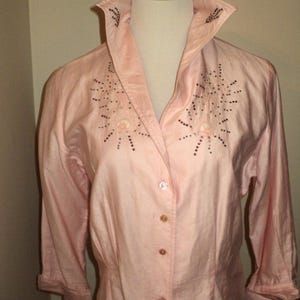 Pu&ograve; includere: Una camicia rosa chiaro con bottoni, colletto ampio e maniche a tre quarti. Il colletto e la parte anteriore della camicia sono impreziositi da perline marrone scuro a forma di stella. La camicia &egrave; esposta su un manichino.