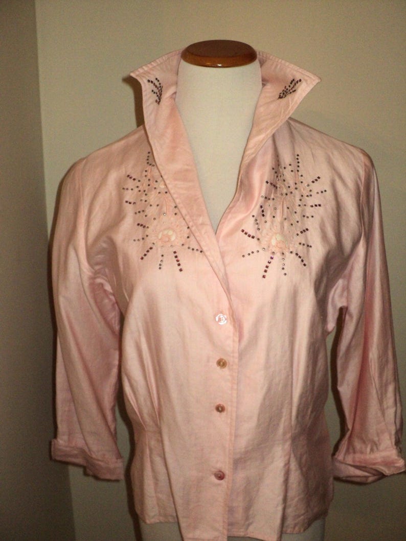 Pu&ograve; includere: Una camicia rosa chiaro con bottoni, colletto ampio e maniche a tre quarti. Il colletto e la parte anteriore della camicia sono impreziositi da perline marrone scuro a forma di stella. La camicia &egrave; esposta su un manichino.