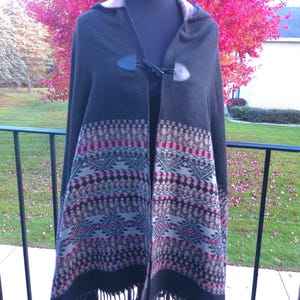 May include: A dark green hooded cape with a patterned design in shades of black, tan, and pink. The cape features a toggle closure and fringe along the bottom edge. The cape is displayed on a mannequin.