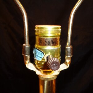 May include: Close-up of a brass lamp socket with a brown knob and the brand name "Stiffel" visible. The lamp socket is attached to a brass pole with two curved brass arms extending upwards.