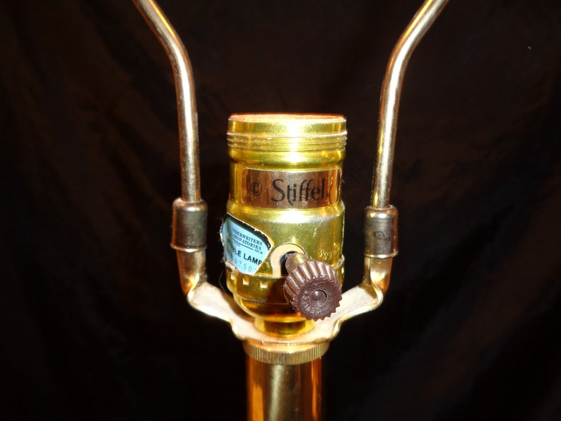 May include: Close-up of a brass lamp socket with a brown knob and the brand name "Stiffel" visible. The lamp socket is attached to a brass pole with two curved brass arms extending upwards.