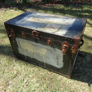 May include: An antique green trunk with metal hardware and corner protectors. The trunk has a rectangular shape with a slightly domed lid. The front of the trunk has a white painted area with text. The trunk is sitting on grass.