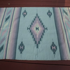May include: A light blue woven rug with a geometric pattern in shades of pink, purple, and green. The rug has a fringed edge.
