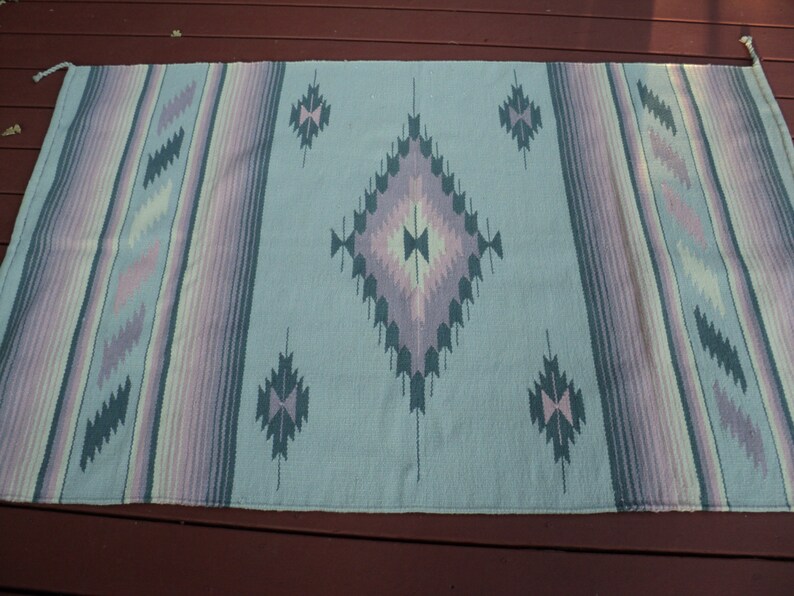 May include: A light blue woven rug with a geometric pattern in shades of pink, purple, and green. The rug has a fringed edge.