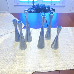 May include: A collection of seven silver-colored metal candle holders with a flared base and a narrow neck. They are arranged on a cream-colored textured table runner, creating a decorative display.