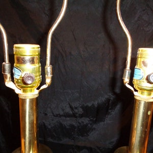 May include: Two brass-colored table lamp bases with curved arms. The lamp sockets are gold-colored with a brown knob and a blue and white label that reads "Stiffel". The lamps are on a black background.