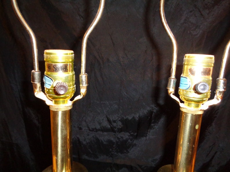 May include: Two brass-colored table lamp bases with curved arms. The lamp sockets are gold-colored with a brown knob and a blue and white label that reads "Stiffel". The lamps are on a black background.
