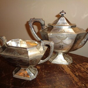 May include: A silver tea set, including a teapot and a sugar bowl, is displayed. The teapot has a lid and a curved spout. Both pieces have ornate detailing and are placed on a wooden surface.