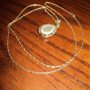 May include: A gold-colored necklace with a teardrop-shaped pendant. The pendant has a light-colored stone set in a decorative gold frame. The chain is a delicate, twisted design, and the necklace is displayed on a wooden surface.