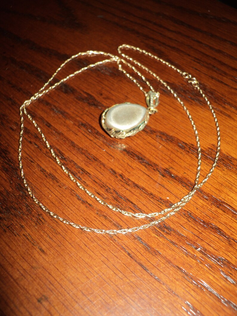 May include: A gold-colored necklace with a teardrop-shaped pendant. The pendant has a light-colored stone set in a decorative gold frame. The chain is a delicate, twisted design, and the necklace is displayed on a wooden surface.