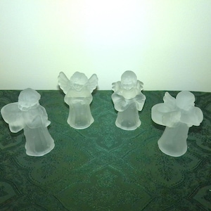 May include: A set of four frosted glass angel figurines standing in a row. Each angel features a translucent, matte finish with detailed wings and robes. The figures are arranged side by side, displaying a consistent sculptural design and form.