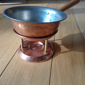 May include: A copper fondue pot with a wooden handle and a brass stand. The pot is sitting on a wooden surface.