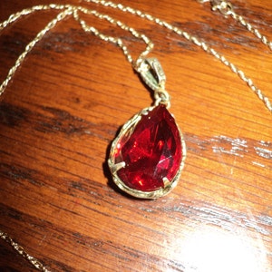 May include: A gold-colored necklace with a teardrop-shaped, faceted red pendant. The pendant is set in a gold-colored frame and hangs from a delicate gold chain. The necklace rests on a wooden surface.