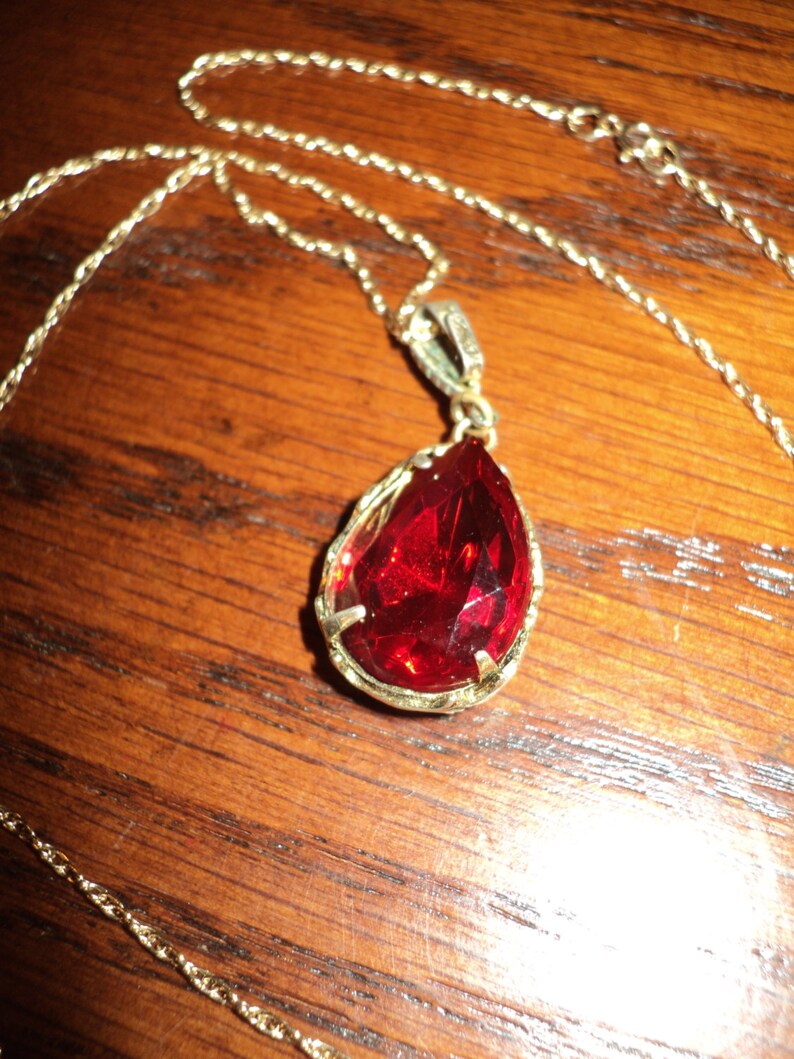 May include: A gold-colored necklace with a teardrop-shaped, faceted red pendant. The pendant is set in a gold-colored frame and hangs from a delicate gold chain. The necklace rests on a wooden surface.