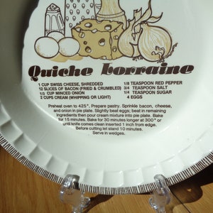 May include: White ceramic plate with a brown border and a recipe for Quiche Lorraine. The recipe includes ingredients like Swiss cheese, bacon, onion, cream, and eggs. The plate is decorated with illustrations of the ingredients.