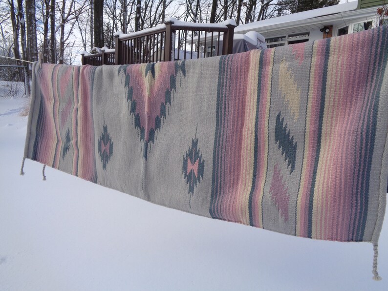 May include: A gray, pink, and yellow striped woven rug with a geometric pattern hangs on a clothesline in the snow.