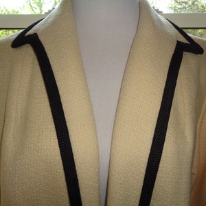 Pode incluir: Um blazer cor de creme com uma guarni&ccedil;&atilde;o preta em volta do colarinho e na frente.