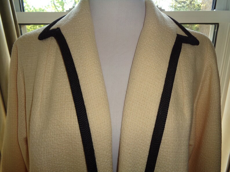 Pode incluir: Um blazer cor de creme com uma guarni&ccedil;&atilde;o preta em volta do colarinho e na frente.
