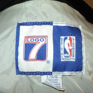 K&ouml;nnte beinhalten: Ein wei&szlig;es und blaues gewebtes Etikett mit dem Text "LOGO 7" in Rot und Wei&szlig; und "OFFICIAL NBA LICENSED PRODUCTO" in Wei&szlig;. Das Etikett hat auch ein kleines eingetragenes Warenzeichen. Das Etikett ist an einem wei&szlig;en Kleidungsst&uuml;ck befestigt.