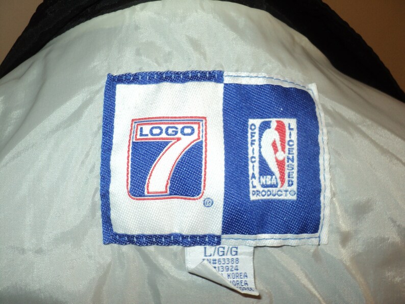 K&ouml;nnte beinhalten: Ein wei&szlig;es und blaues gewebtes Etikett mit dem Text "LOGO 7" in Rot und Wei&szlig; und "OFFICIAL NBA LICENSED PRODUCTO" in Wei&szlig;. Das Etikett hat auch ein kleines eingetragenes Warenzeichen. Das Etikett ist an einem wei&szlig;en Kleidungsst&uuml;ck befestigt.