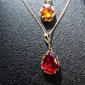 May include: Two gold-toned necklaces. The top necklace features a round, amber-colored pendant with a floral design. The bottom necklace has a teardrop-shaped, faceted, red pendant. Both necklaces are on a black surface.