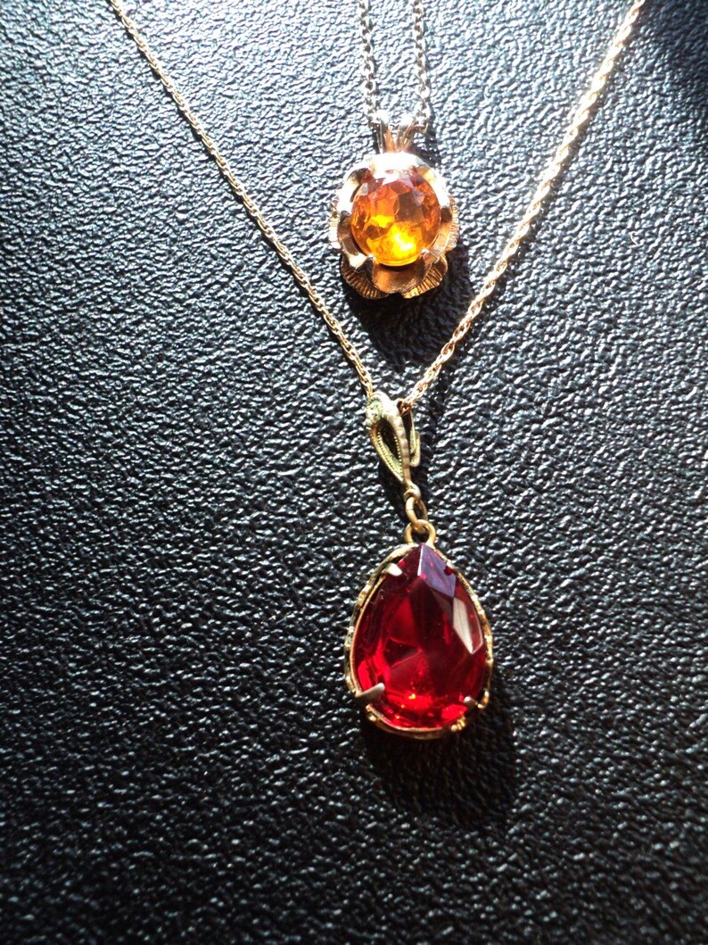 May include: Two gold-toned necklaces. The top necklace features a round, amber-colored pendant with a floral design. The bottom necklace has a teardrop-shaped, faceted, red pendant. Both necklaces are on a black surface.