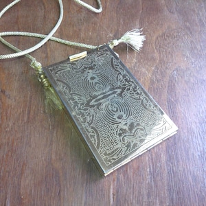 May include: A rectangular, gold-colored metal purse with an ornate, engraved design. The purse has a matching gold cord strap and tassel. The clasp is also gold. The purse is lying on a wooden surface.