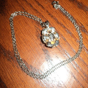 May include: Silver-toned necklace with a floral pendant. The pendant features a flower design with white and gold accents. The chain is a delicate link style, and the necklace is displayed on a wooden surface.