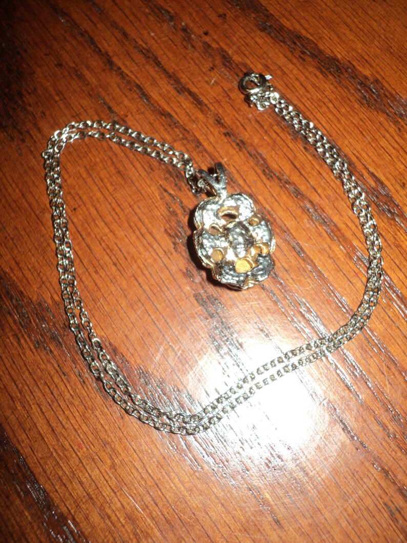 May include: Silver-toned necklace with a floral pendant. The pendant features a flower design with white and gold accents. The chain is a delicate link style, and the necklace is displayed on a wooden surface.