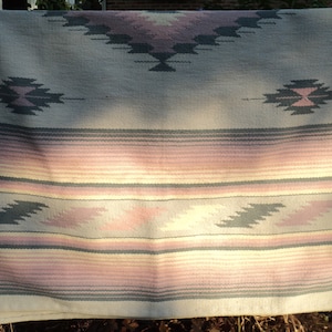 May include: A woven throw blanket with a Southwestern design. The blanket is made of a soft, light-colored fabric with a geometric pattern in shades of pink, gray, and white.