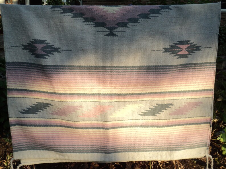 May include: A woven throw blanket with a Southwestern design. The blanket is made of a soft, light-colored fabric with a geometric pattern in shades of pink, gray, and white.