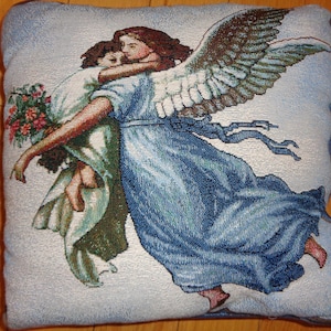 Op de afbeelding: Vierkant kussen met een geweven tapijtontwerp. De afbeelding toont twee figuren die elkaar omarmen, een met grote vleugels, in tinten blauw, groen en bruin. De figuren houden een boeket bloemen vast. Het kussen heeft een lichtblauwe achtergrond.