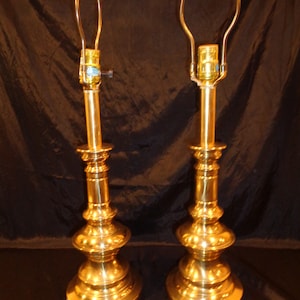 May include: Two vintage brass table lamps with a classic, ornate design. Each lamp features a tiered base, a slender central column, and a curved harp for holding a lampshade. The lamps are polished to a high shine, reflecting the light.