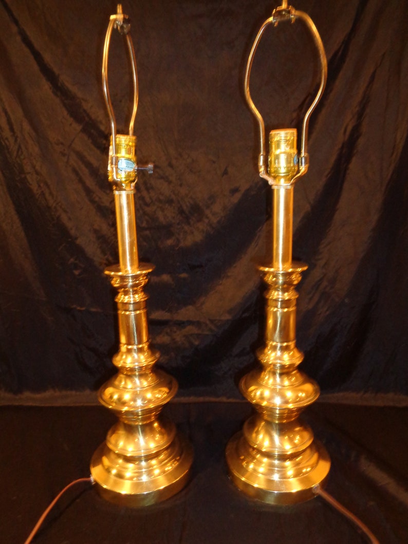 May include: Two vintage brass table lamps with a classic, ornate design. Each lamp features a tiered base, a slender central column, and a curved harp for holding a lampshade. The lamps are polished to a high shine, reflecting the light.