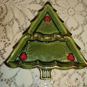 May include: A green ceramic Christmas tree-shaped serving dish with gold trim. The dish features red berry accents and a detailed design. The tree-shaped dish is a festive addition to holiday decor.