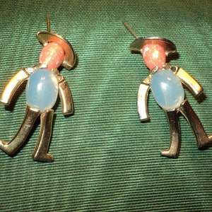 May include: A pair of gold-tone earrings featuring a whimsical design of a person with a pink hat and a light blue body. The earrings are made of metal and have a vintage style.