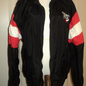 K&ouml;nnte beinhalten: Schwarze Windbreaker-Jacke mit roten und wei&szlig;en Streifen an den &Auml;rmeln. Die Jacke hat einen Rei&szlig;verschluss und ein Logo mit der Aufschrift "Chicago Bulls" auf der rechten Seite.