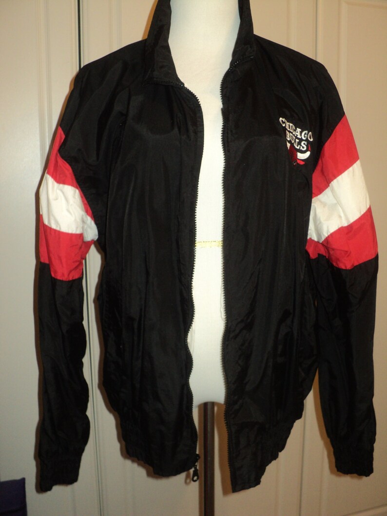 K&ouml;nnte beinhalten: Schwarze Windbreaker-Jacke mit roten und wei&szlig;en Streifen an den &Auml;rmeln. Die Jacke hat einen Rei&szlig;verschluss und ein Logo mit der Aufschrift "Chicago Bulls" auf der rechten Seite.