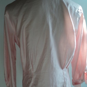 Pu&ograve; includere: Una camicia rosa chiaro a maniche lunghe con colletto classico e polsini abbottonati. La parte posteriore della camicia presenta un design su misura con cuciture verticali e vita aderente. Il tessuto sembra liscio, forse seta o raso.