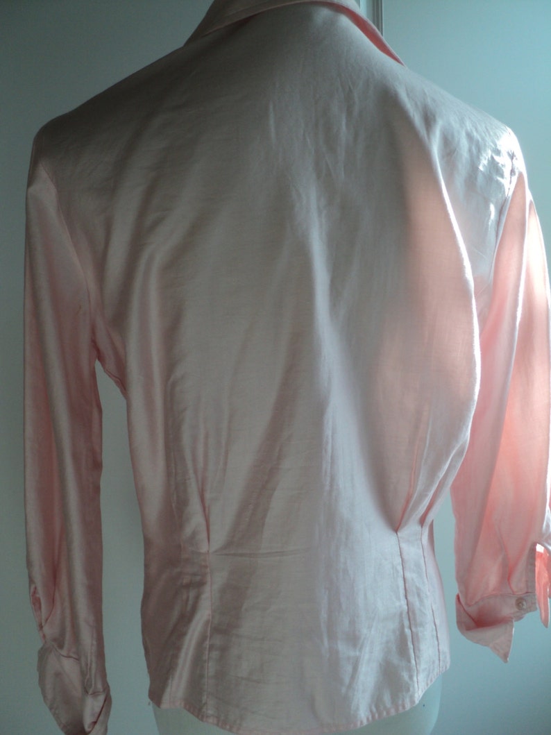 Pu&ograve; includere: Una camicia rosa chiaro a maniche lunghe con colletto classico e polsini abbottonati. La parte posteriore della camicia presenta un design su misura con cuciture verticali e vita aderente. Il tessuto sembra liscio, forse seta o raso.