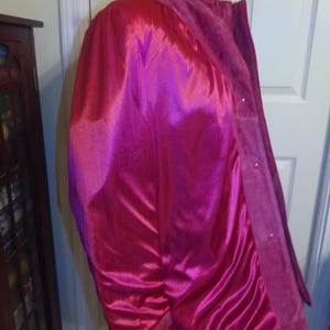 May include: A vibrant pink coat with a high collar and button closure. The coat features a satin-like lining and a suede-like exterior, creating a contrast in textures. The garment is displayed on a wooden hanger.