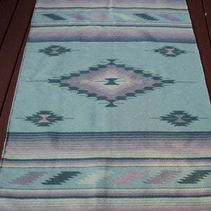 May include: A light blue woven rug with a geometric pattern in shades of purple, pink, and green. The rug has a fringed edge.