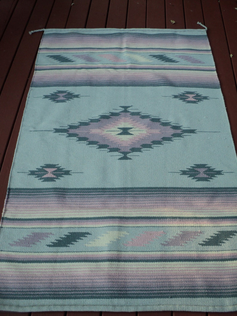 May include: A light blue woven rug with a geometric pattern in shades of purple, pink, and green. The rug has a fringed edge.